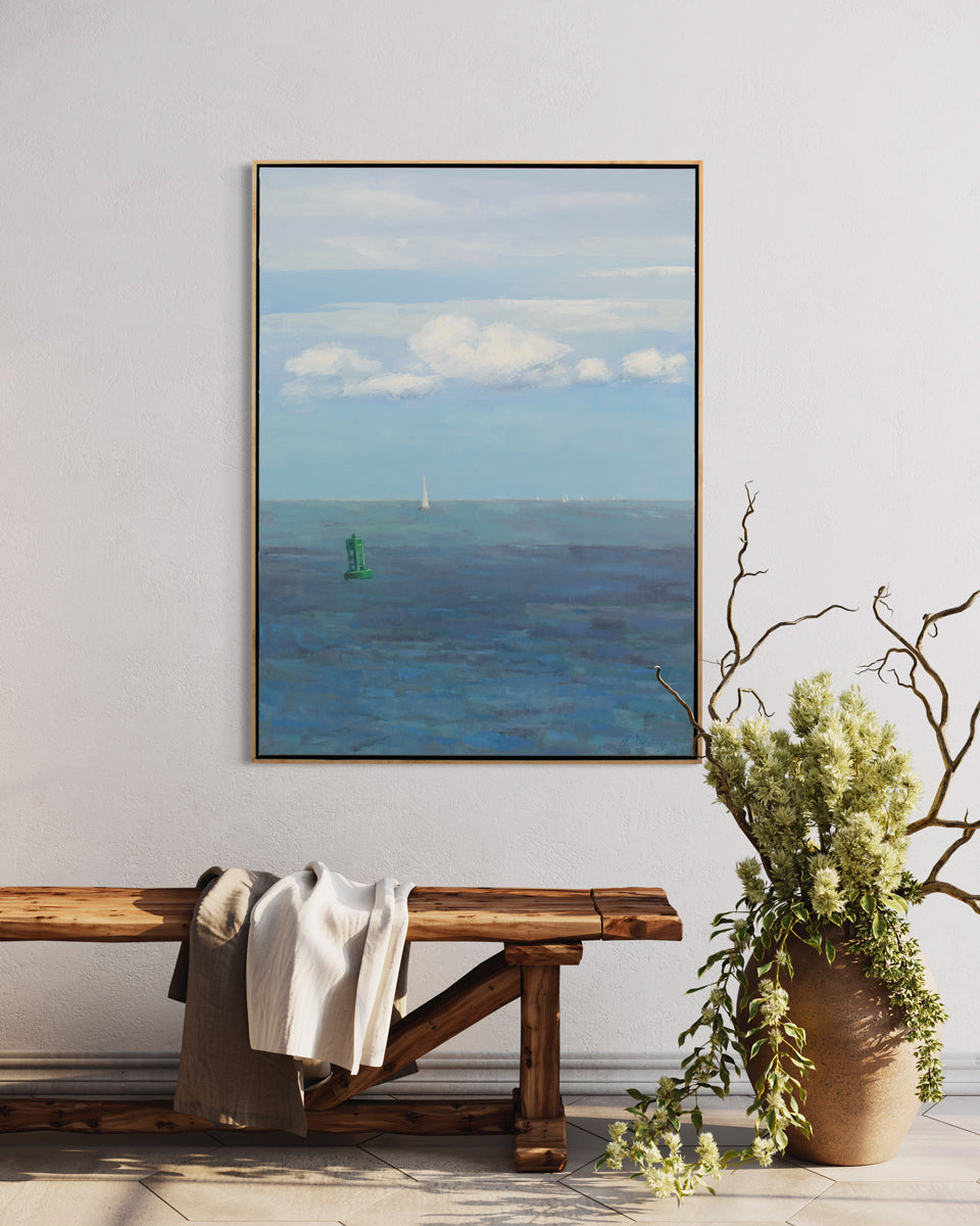 A blue, white and green coastal landscape painting by Molly Doe Wensberg hangs in a light wood floater frame on a white wall above a carved wood bench with clothing laying over and a potted topiary.