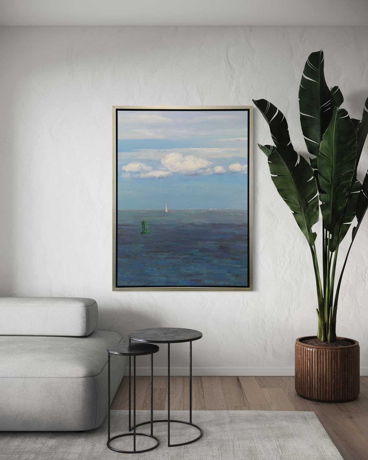 A coastal seascape print in a silver floater frame hangs on a white wall above the end of a white modern sofa, two black side tables and a tall potted floor plant.