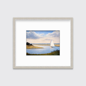 A print of a sailboat going down a winding blue river in a silver frame and mat hangs on a white wall.