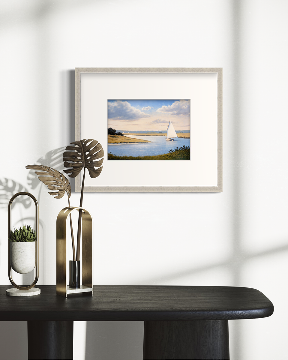 A print of a sailboat sailing though a bay in a silver frame hangs over a black table with a small plant and a decorative accessory.