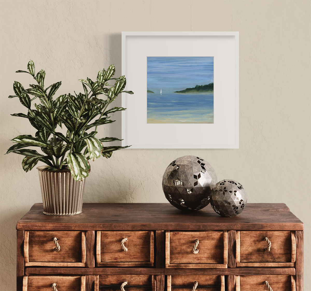 A blue, green and white abstract seascape print with a small sailboat in a white frame with a mat hangs on an off white wall above a natural wood drawer cabinet with a potted plant and two silver spheres.