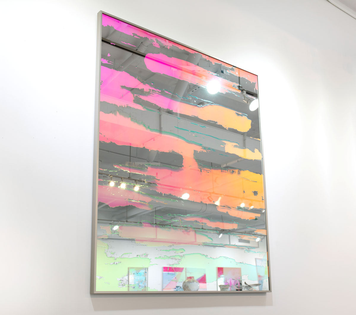 A pink fine art mirror hangs on a white wall in an art gallery.