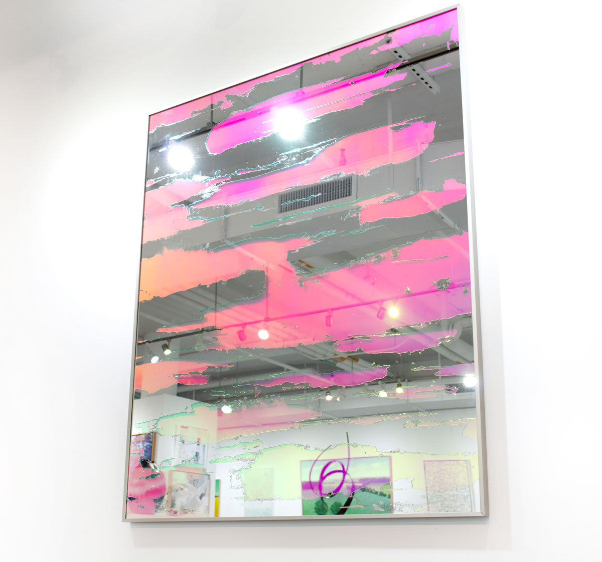 A pink fine art mirror hangs on a white wall in an art gallery. 