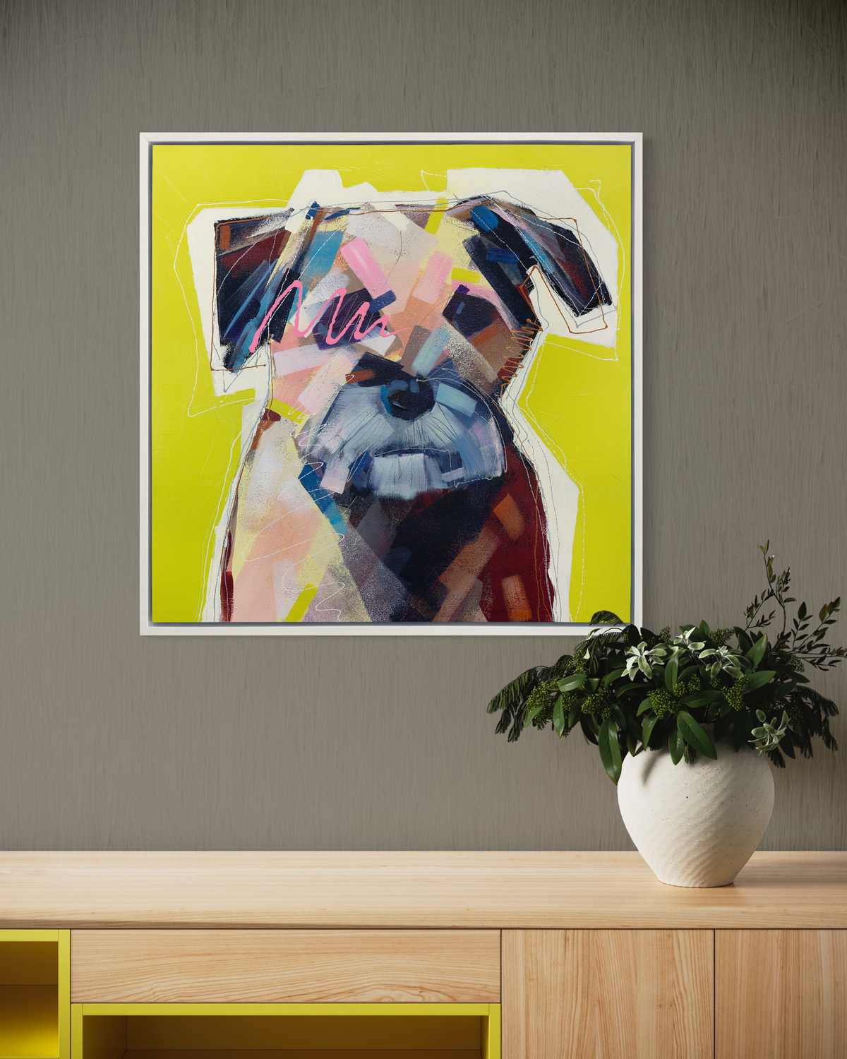 A multicolored abstract dog print in a white floater frame hangs on a green-grey wall above a wood shelf with a potted plant.