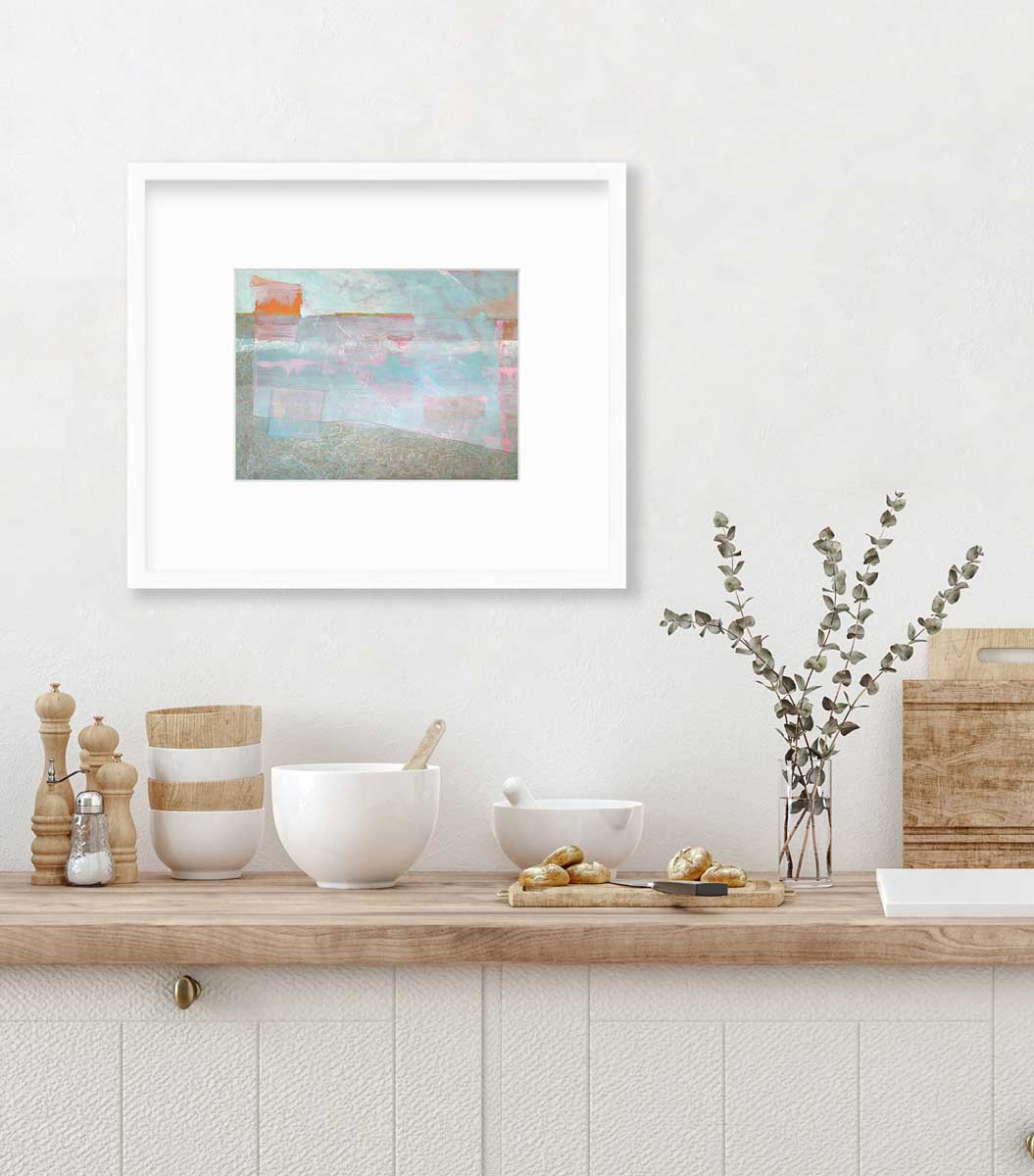 A light blue and pink abstract print in a white frame with a mat hangs on a white wall above a butcher block countertop with bowls, a cutting boards and a clear vase with eucalyptus stems.