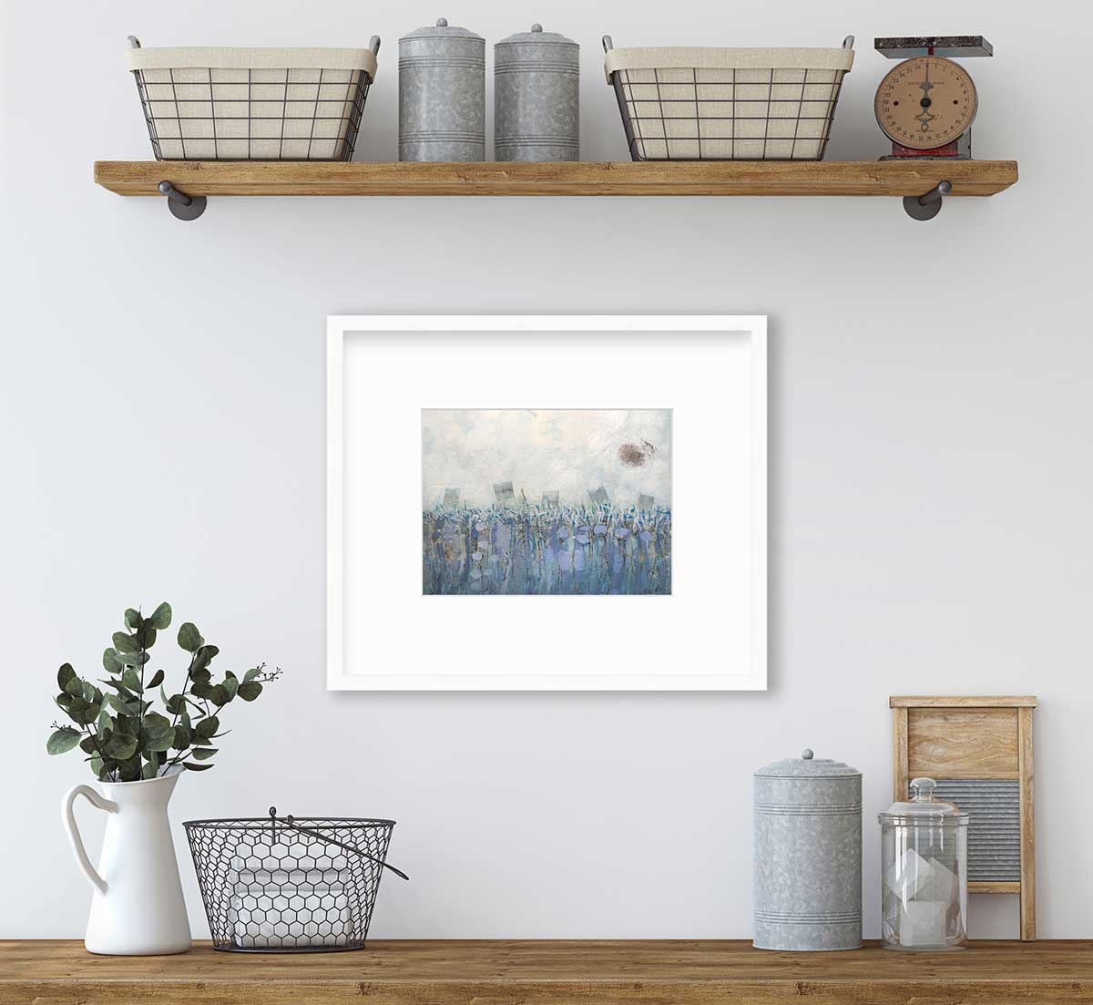 A beige, teal and lavender abstract print in a white frame with a mat hangs on a white wall above a wood surface with a white pitcher with eucalyptus leaves, a silver container and a black wire basket.