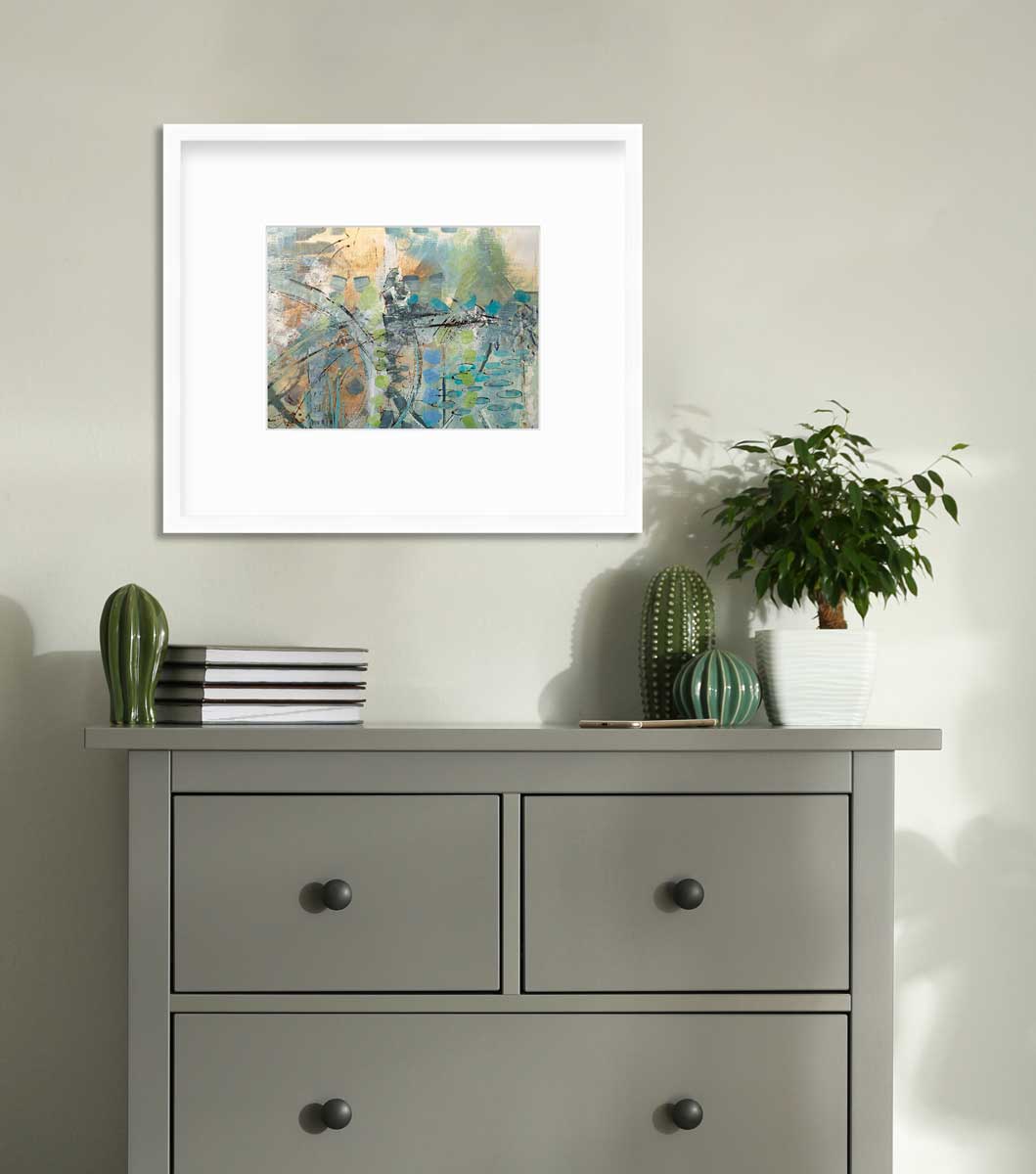 A blue, green, orange and black abstract print in a white frame with a mat hangs on a light green wall above a grey dresser with a stack of books, three green cacti decorations and a small potted tree.