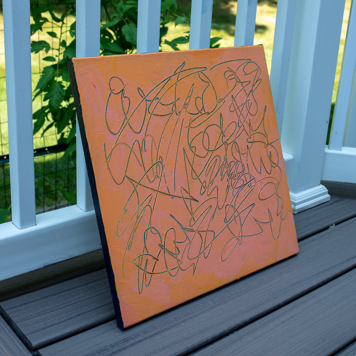 An abstract text based painting with a peach-orange background and teal carved scribbled line art, leaning against a white railing on a gray deck with green grass in the background.