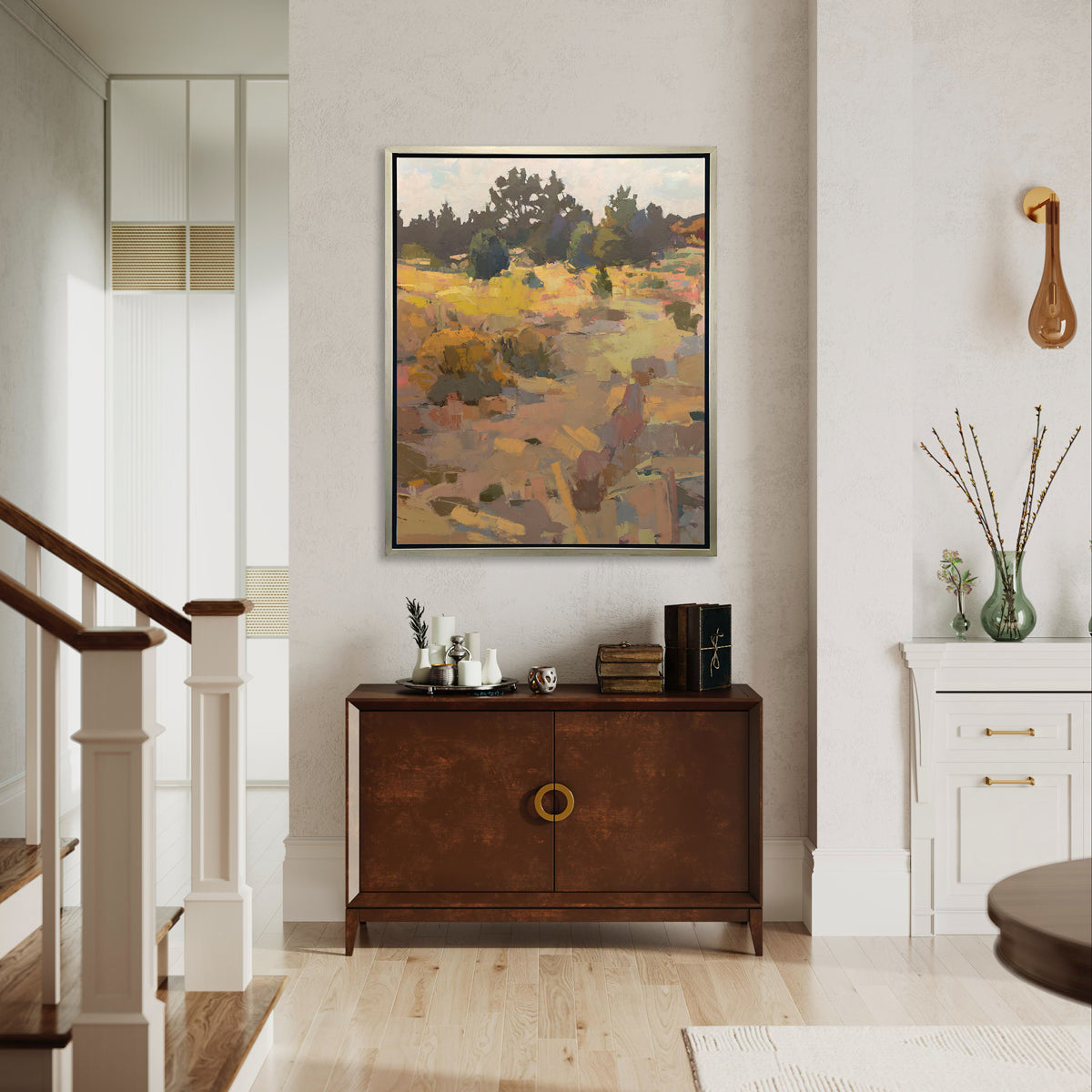 An earth-toned abstracted landscape art print framed in a silver frame hangs on a white wall above a wooden console table with vintage table decor next to the bottom of a white and wood staircase. 