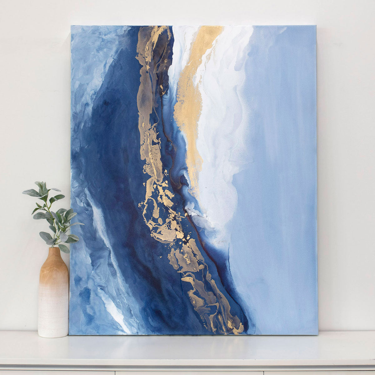 Abstract art painting with blue and gold tones on a white wall, accompanied by a vase with greenery.