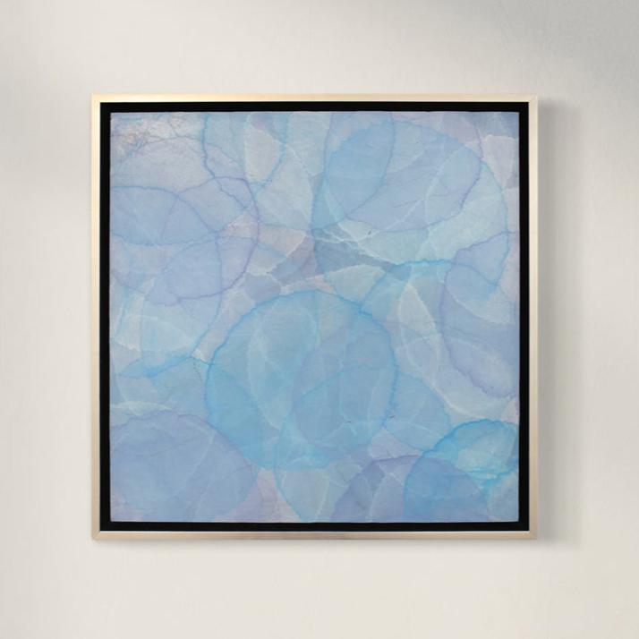 A tonal blue layered circle abstract painting in a silver floater frame hangs on a white wall.