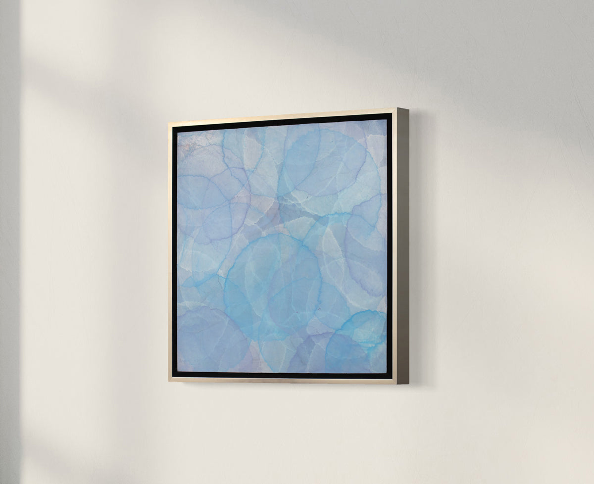 A side view of a tonal blue layered circle abstract painting in a silver floater frame hangs on a white wall.