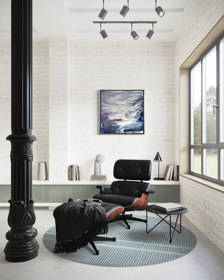 A blue, white, and lavender print framed in silver floater frame hangs on a white wall with a black modern leather chair and side table in front.