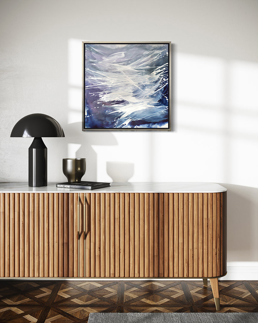 A blue, white, lavender print framed in silver floater frame hangs on a white wall  over a midcentury TV cabinet with a black lamp, vase and book.