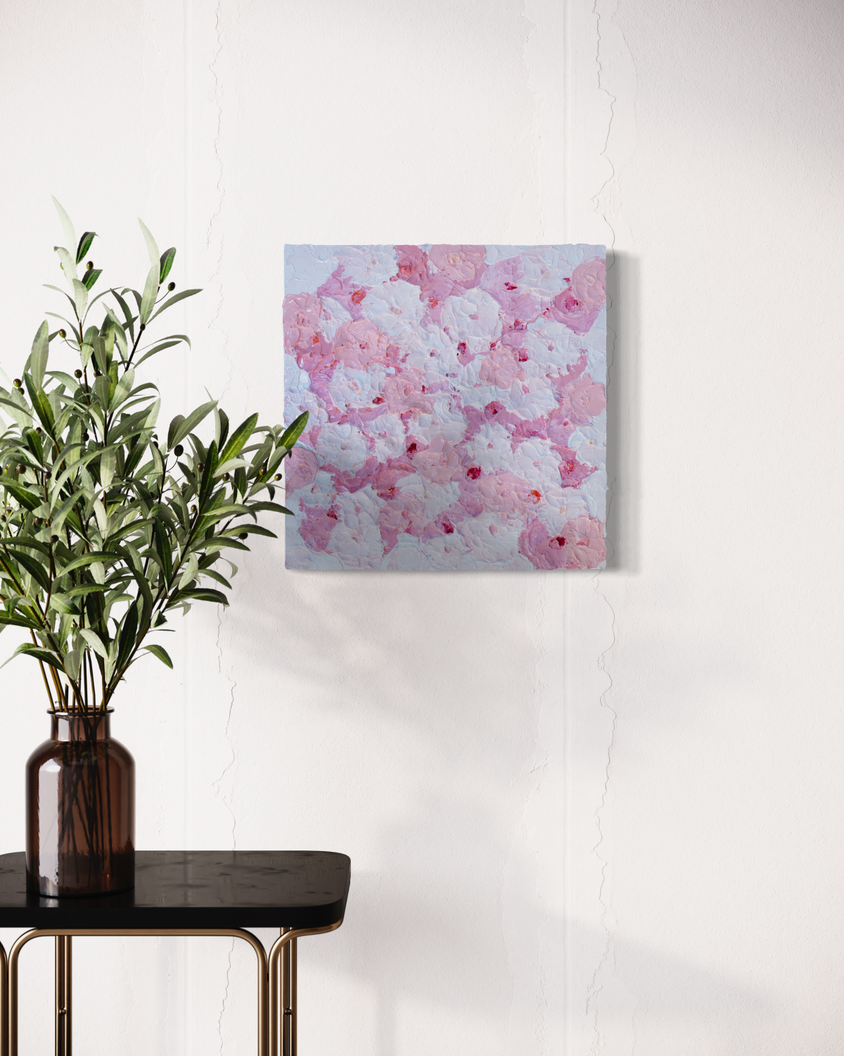 A close up of a very textured abstract painting with circle type abstract flowers in pink and white hangs on a white wall above an end table with a glass vase holding eucalyptus branches.