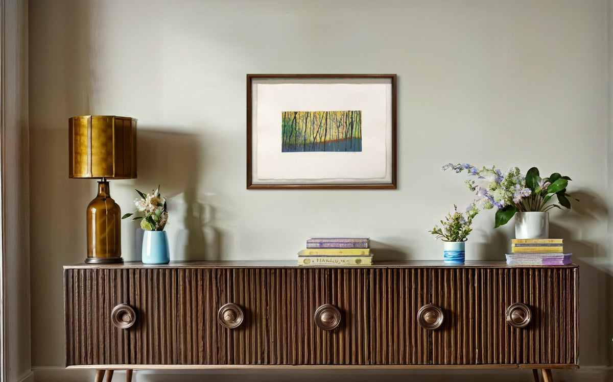 Framed artwork of a forest scene with trees in yellow and green hanging on a white wall over a mid century credenza.