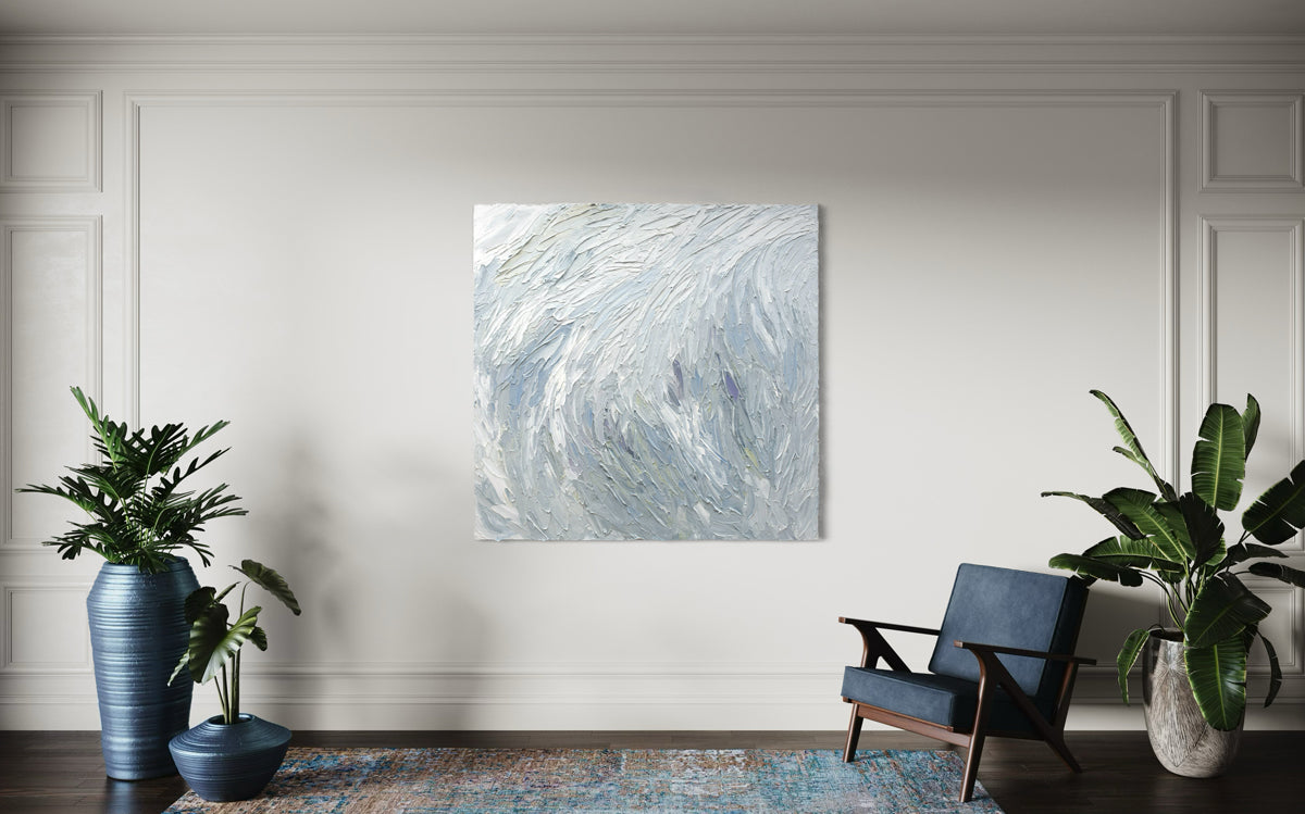 A Grey blue, light blue, lavender, celadon and white thickly textured abstract painting by Teodora Guererra hanging on a white wall in a family room. An oversized blue chair and ottoman sit in front.
