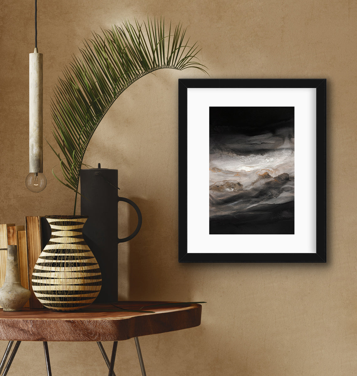 A black, white, beige and gold abstract print in a black frame with a mat hangs on a beige wall next to a live edge side table with a black and beige vase with a giant leaf stem, a black canister and a stack of books.
