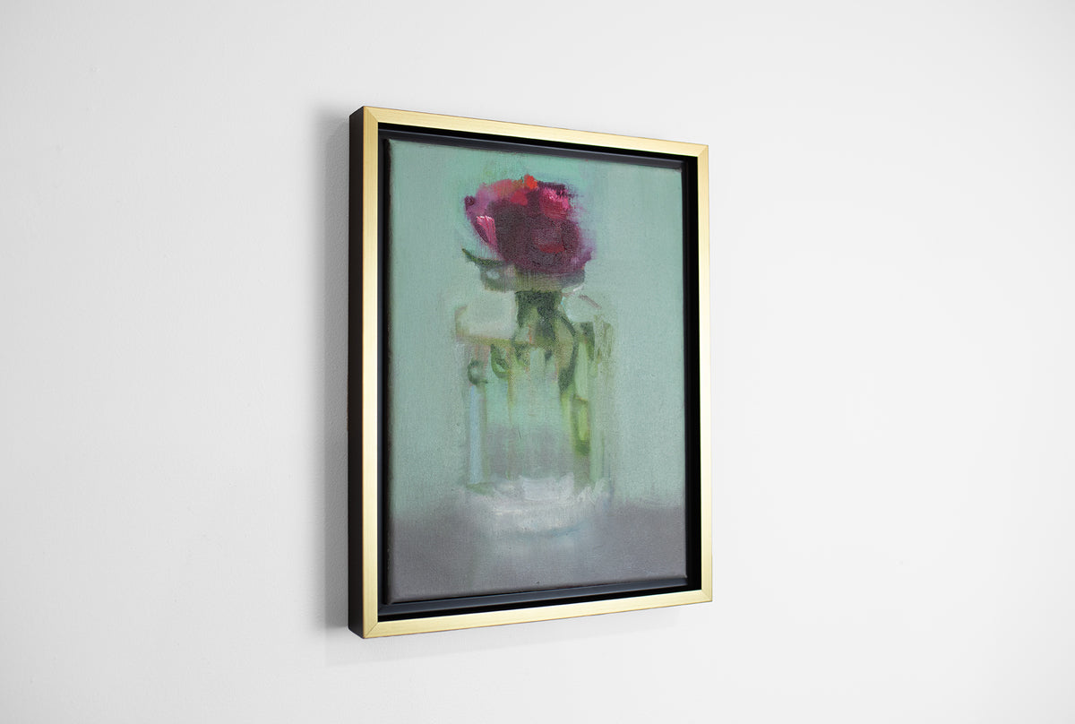 A small framed painting of a red flower in a clear glass vase, framed in a gold floating frame, hangs on a white wall.