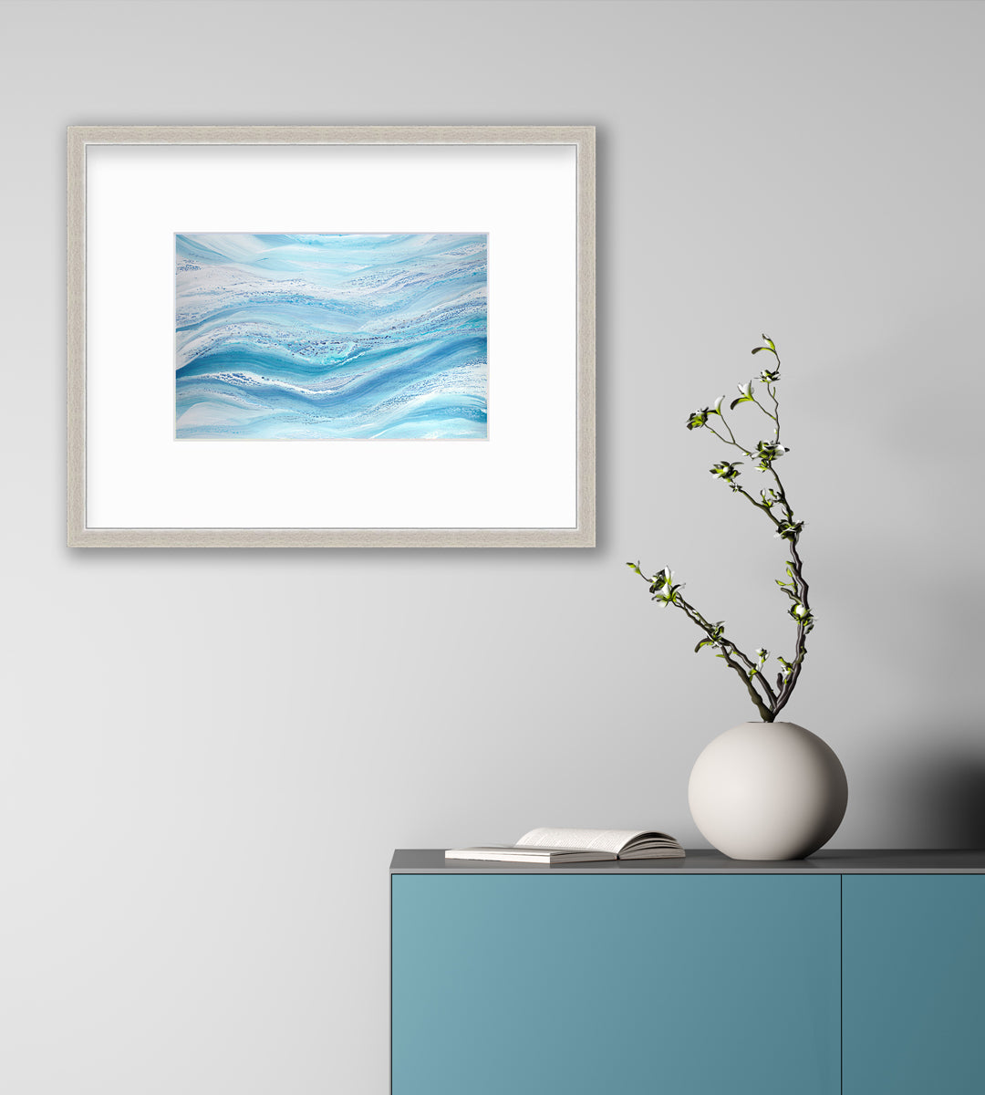 A blue, teal and white abstract print in a silver frame with a mat hangs on a white wall above a teal cabinet with an open book and rounded white vase with greenery stems.