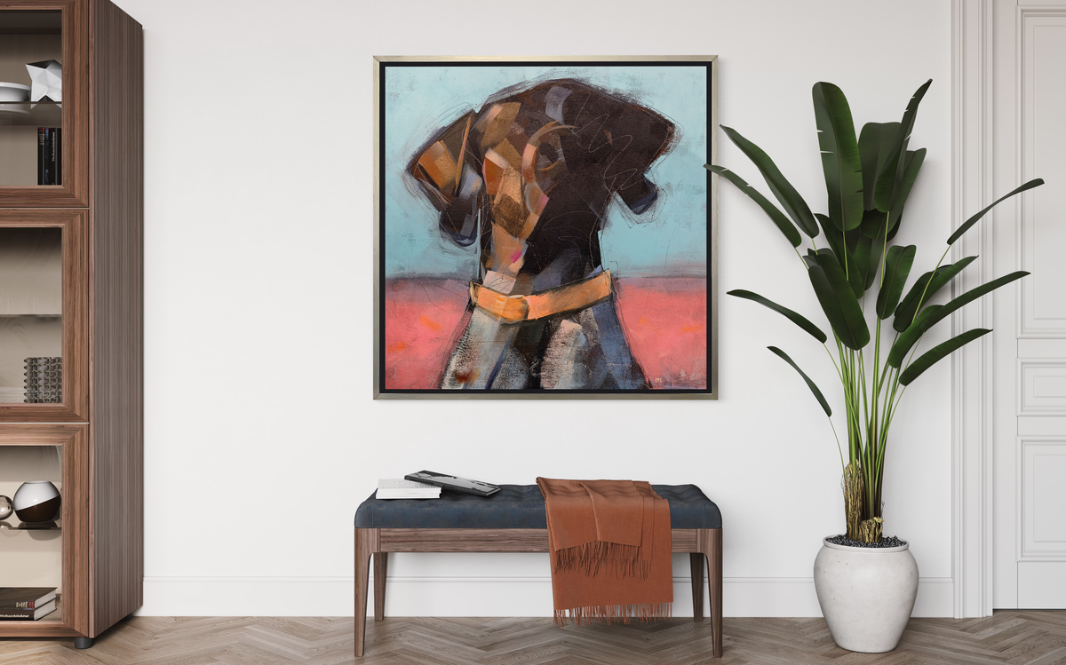 A brown, white, pink and blue abstract dog print in a silver floater frame hangs on a white wall above a wood bench with a denim blue cushion, a dark orange throw blanket and a tall potted plant to the side.