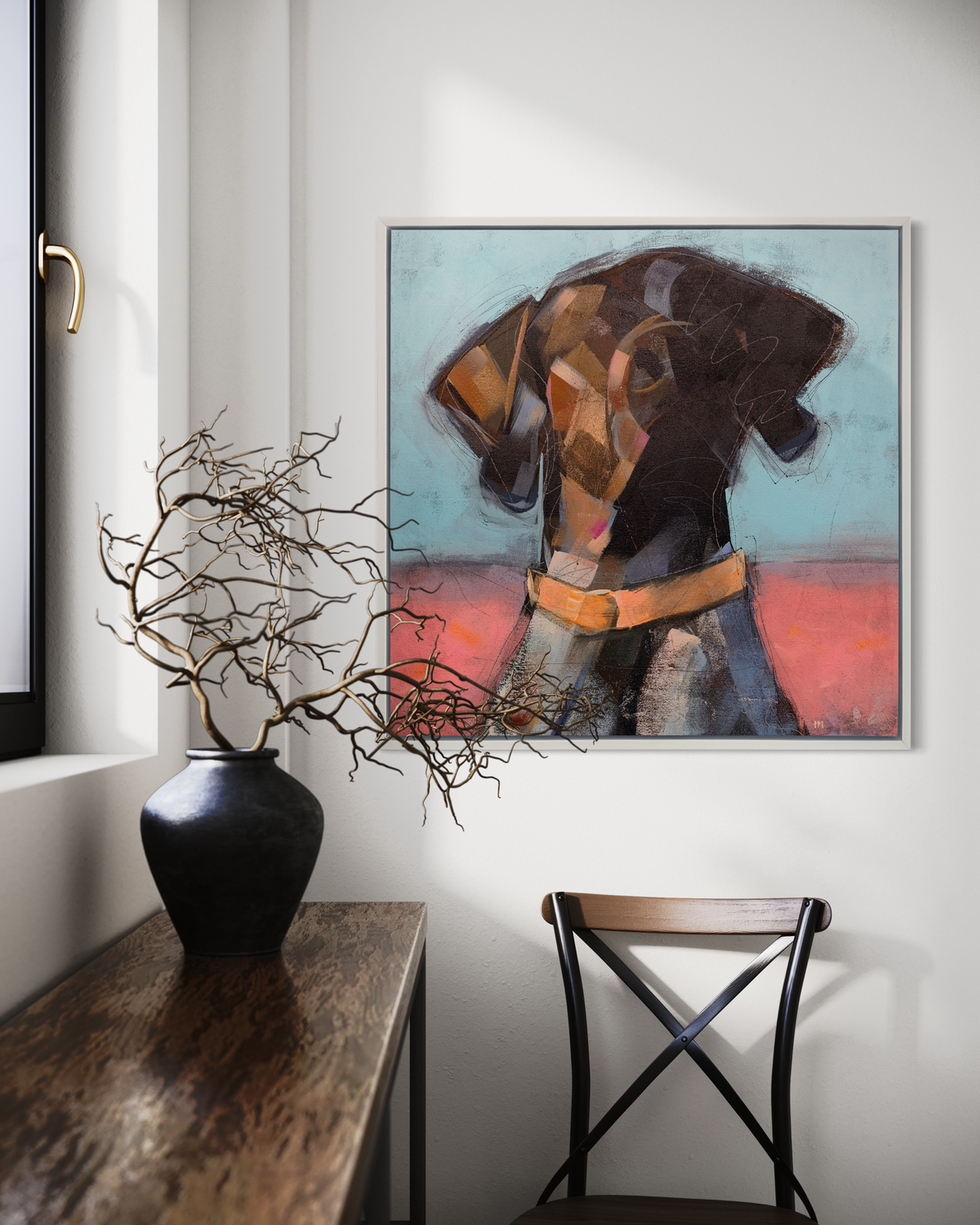 A brown, white, pink and blue abstract dog print in a white floater frame hangs on a white wall above a wood chair and next to a wood console table with a dark vase holding wood stems.