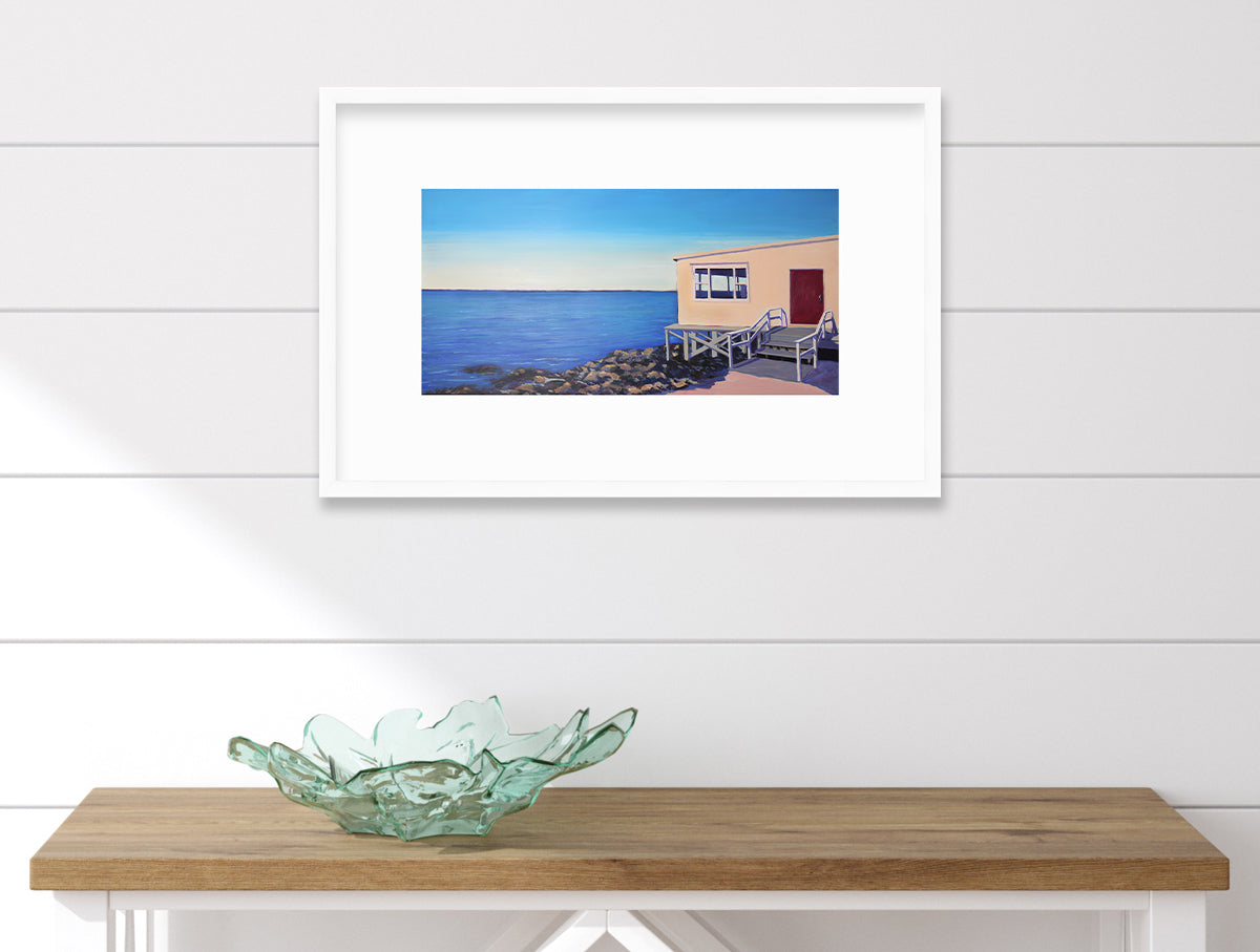 A contemporary seascape print in a white frame with a mat hangs on a white paneled wall above a wood console with white legs and a green glass bowl sitting on top.