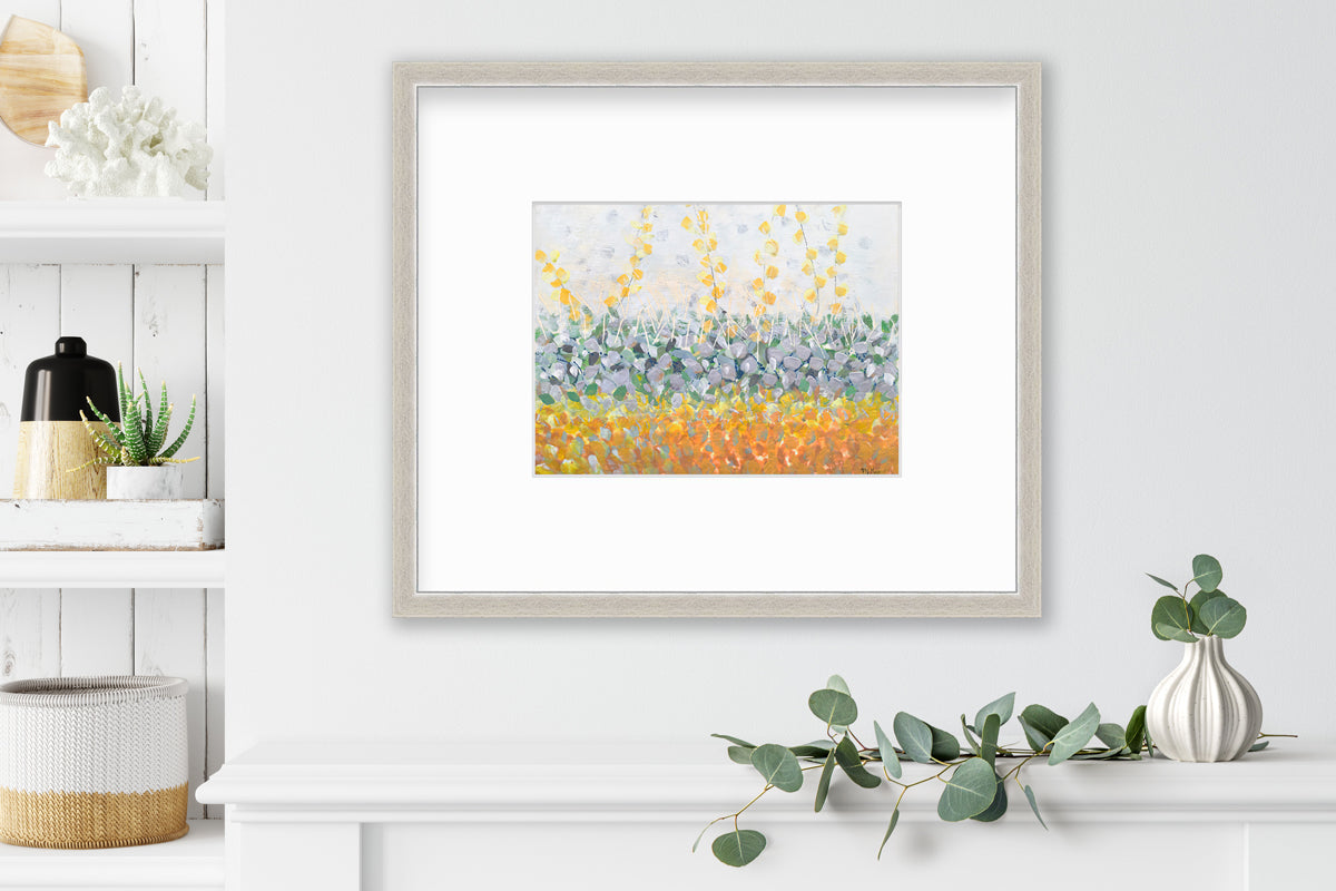 A yellow, purple, green and orange abstract print in a silver frame with a mat hangs on a white wall above a white shelf with a eucalyptus filled white vase and next to a built in bookshelf with decorative accessories.