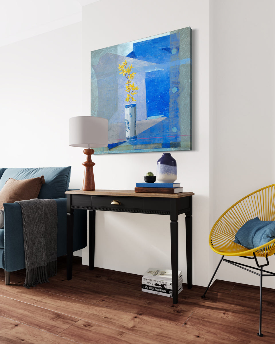 An abstracted blue painting with a vase with yellow flowers in the foreground hangs on a white wall above a black console table holding a table lamp and table decor. It is between a yellow modern seat with a blue throw pillow and a blue sofa on the left. 