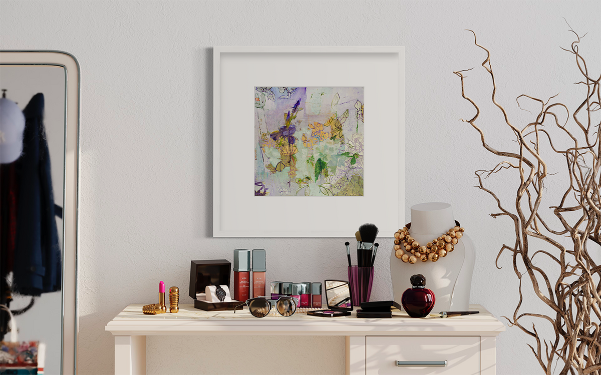A purple, gold and green abstract print in a white frame with a mat hangs on a white wall above a small desk with makeup and jewelry.