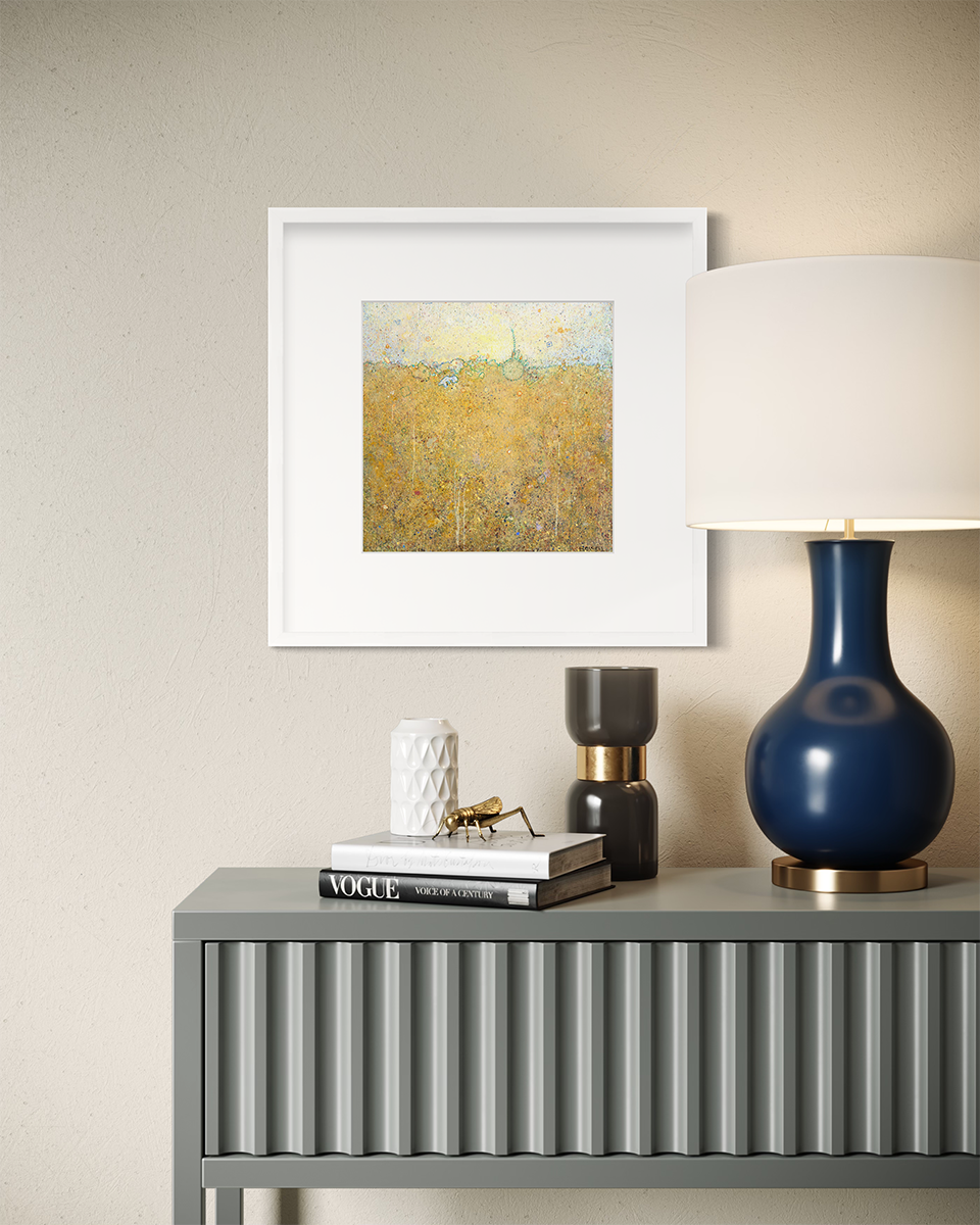 A yellow abstract landscape print in a white frame with a mat hangs on a white wall above a grey console table with a navy blue lamp, a stack of books and a few decorative accessories.