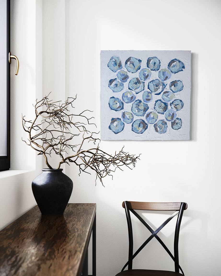 A very textured abstract painting with circle type abstract flowers in  blue, teal, green, plum hangs on a white wall above a chair, A console sits to the left with a pot of dried decorative branches.