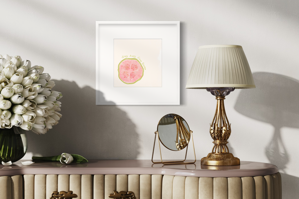 An abstract art print of a fruit with the words "stay away from drama" in a white frame with a mat hangs on a white wall above a blush and beige vanity with a small mirror, an ornate table lamp and a vase full of white tulips.