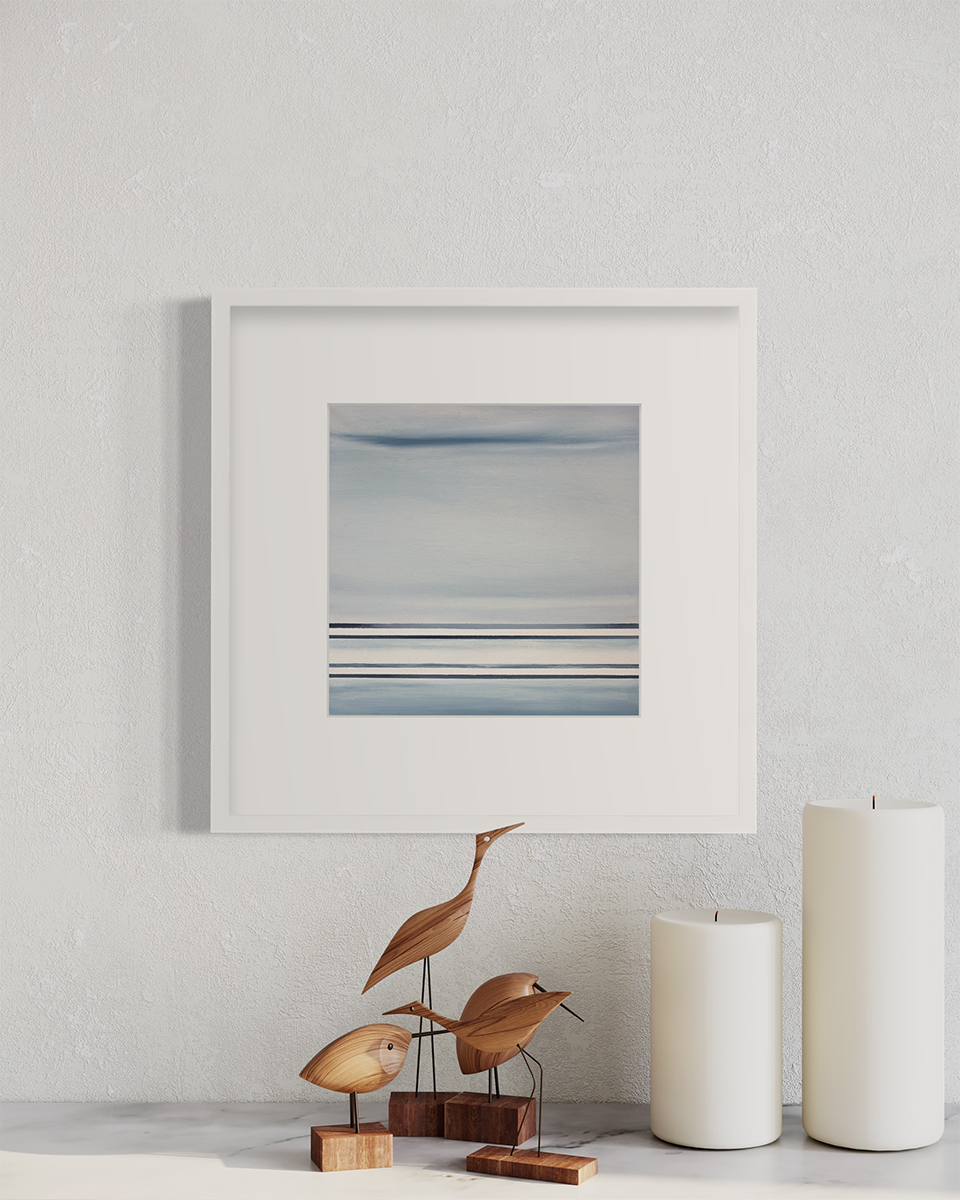 A blue and white linear abstract print in a white frame with a mat on a white wall above two white pillar candles and wooden bird figurines.