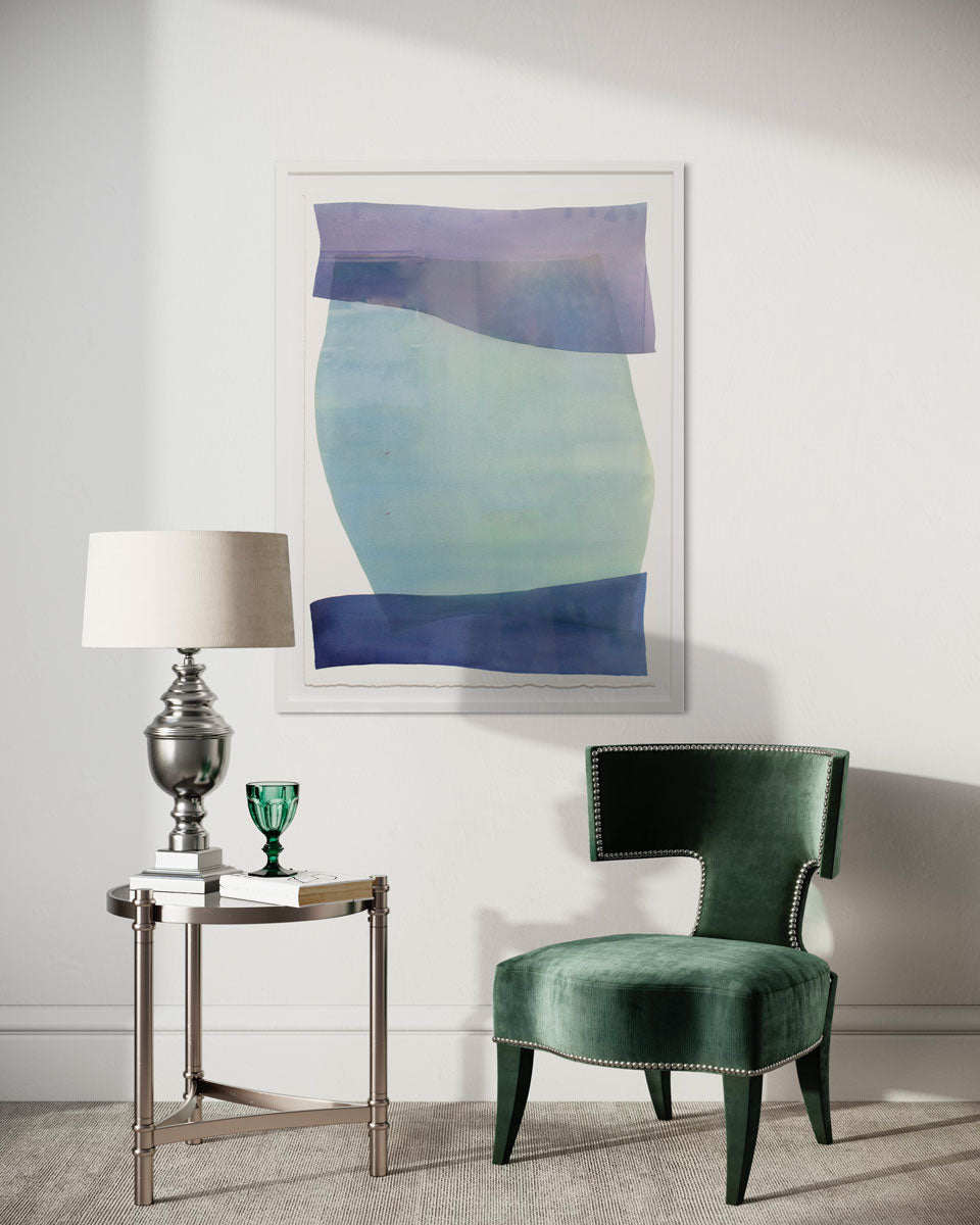 A blue and green abstract watercolor painting framed in a white frame hangs on a white wall above a green velvet chair and a silver end table with a traditional lamp, book, and green glass. 