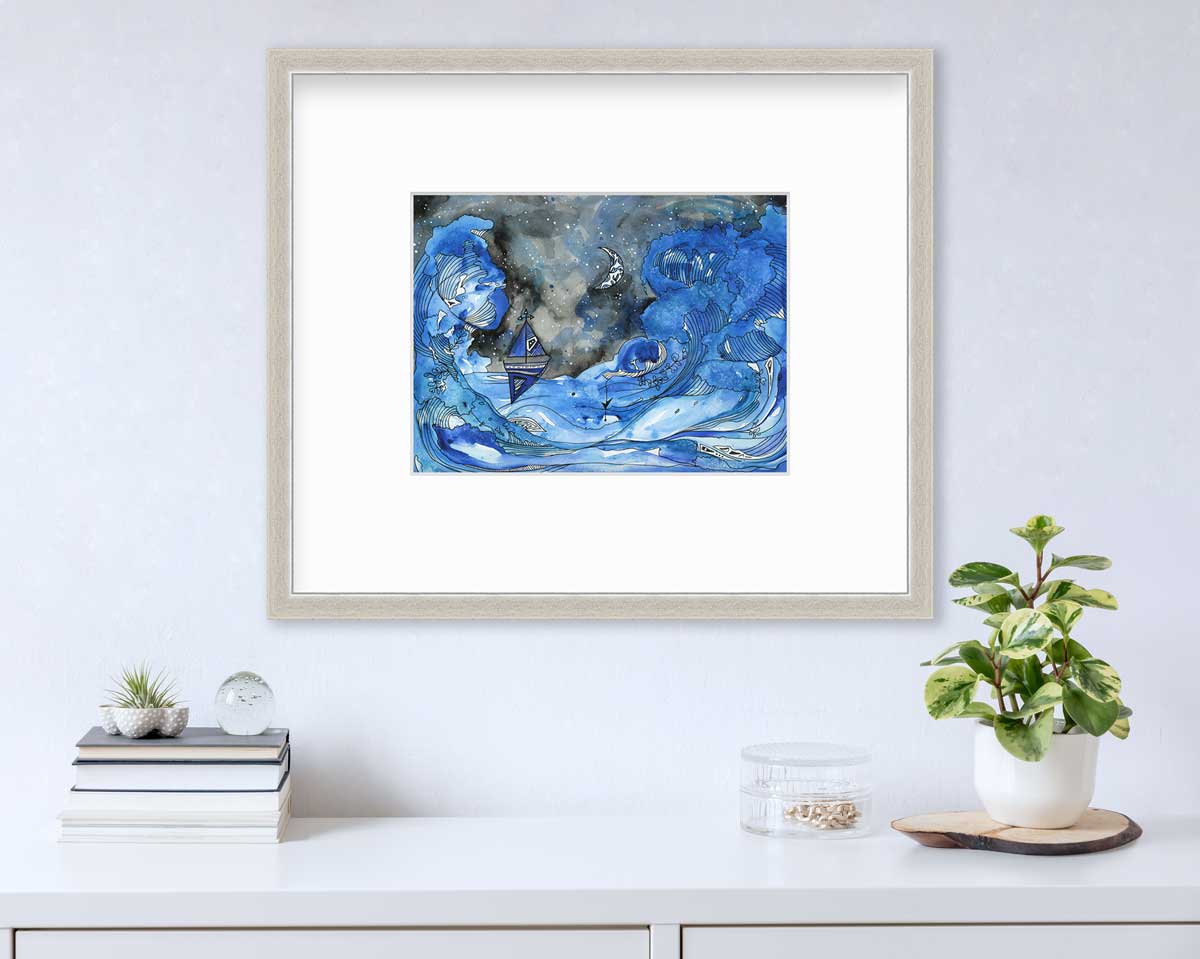A black, blue and white seascape illustration print framed in a silver frame with a mat hangs on a white wall above a white surface with a stack of books, a clear round container with gold accessories and a potted plant.
