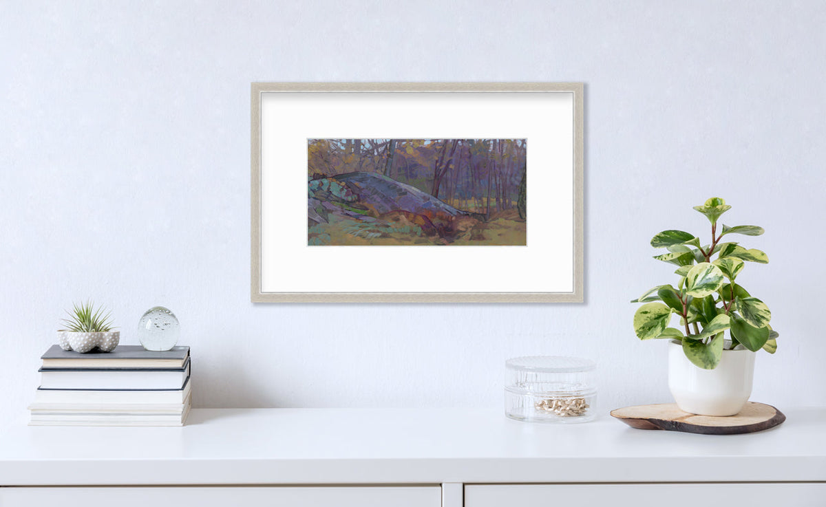 A muted multicolored abstract landscape print in a silver frame with a mat hangs on a white wall above a white surface with a stack of books, a round clear container with gold accessories and a potted plant.