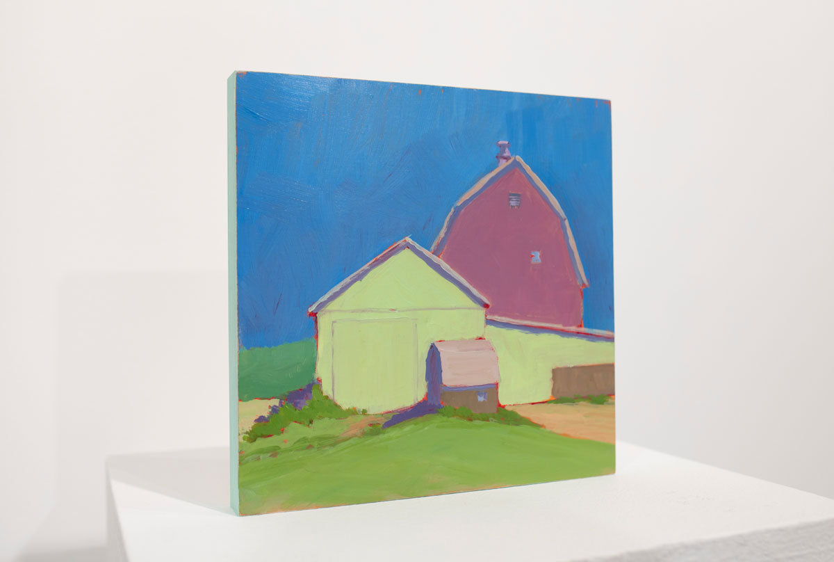 A blue, pink, and green contemporary landscape painting sits upright on a white table surface in front of a white wall.