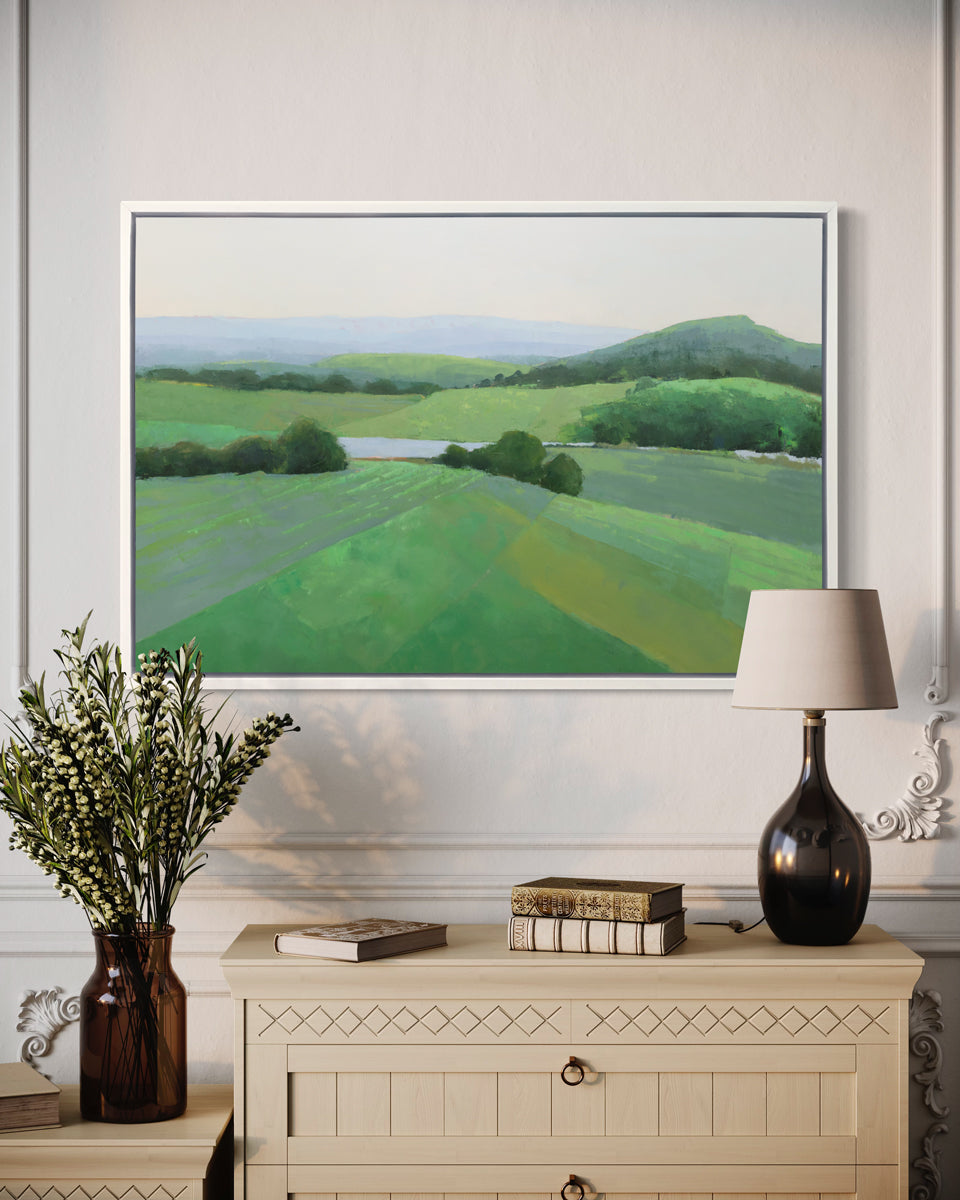 A green landscape art print framed in a white frame hangs on a light wall above a light wood dresser with ornate, traditional table decor. 