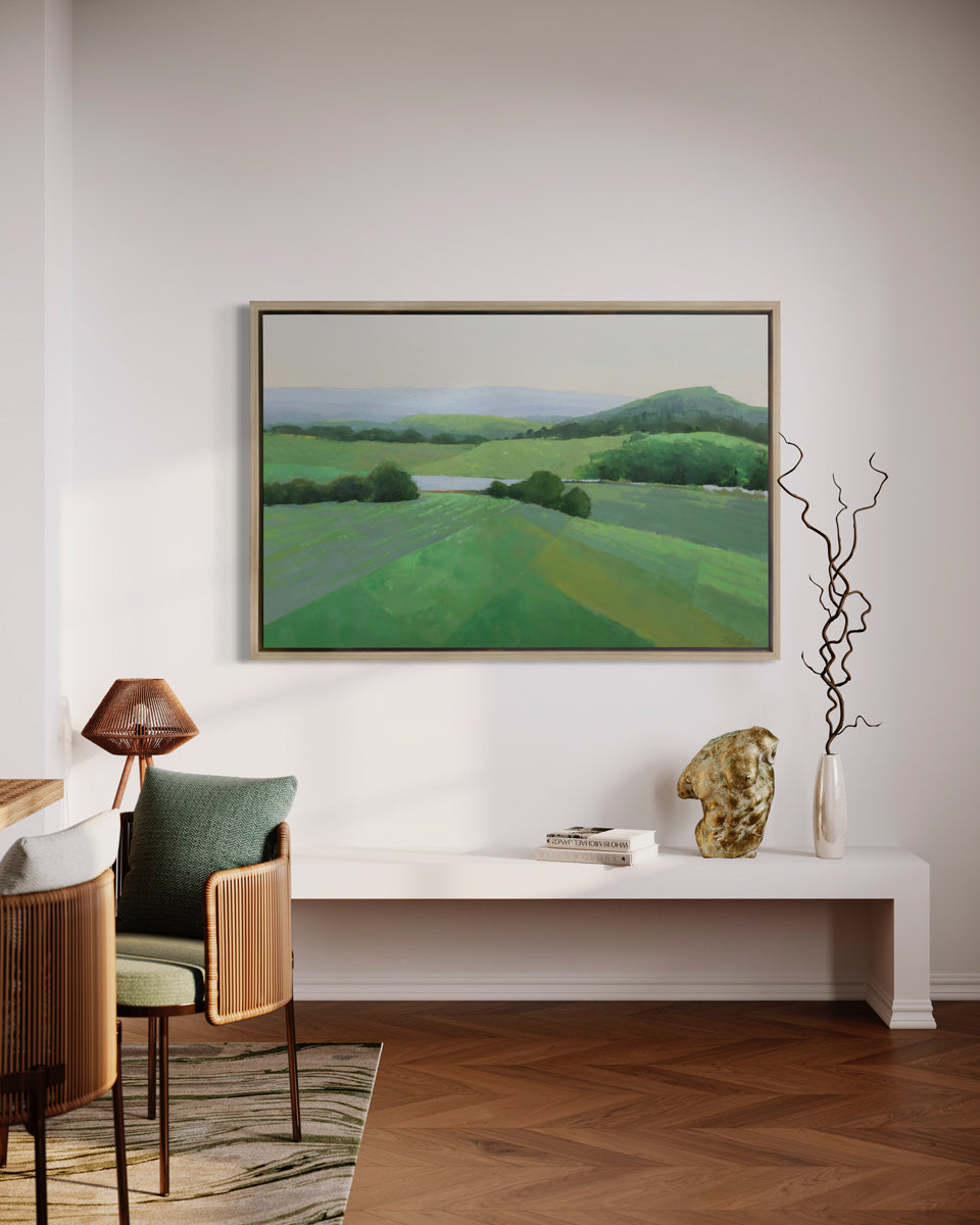 A green landscape art print framed in a white oak frame hangs on a white wall above a modern white bench with modern table decor, and wooden table with chairs. 