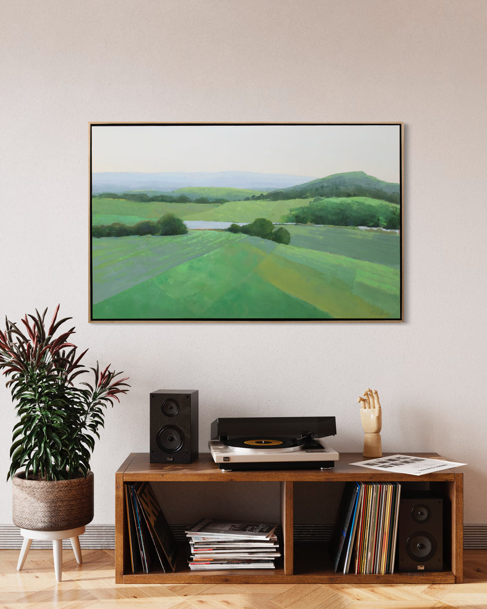 A green landscape painting by Molly Doe Wensberg framed in a wood frame hangs on a light wall above a small wooden shelving unit with records and a record player, and a potted floorplant. 