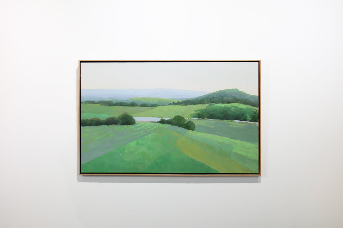 A green landscape painting framed in a light wood floater frame hangs on a white wall.