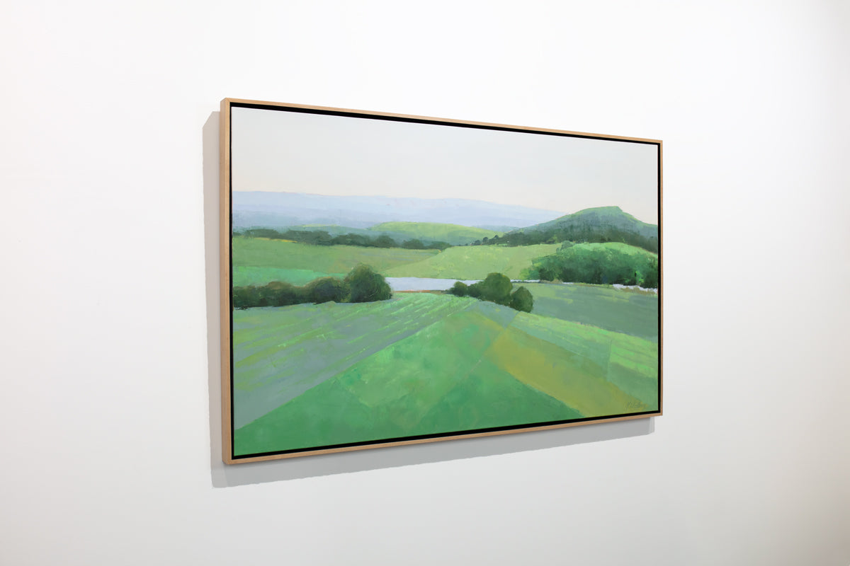 A green landscape painting framed in a light wood floater frame hangs on a white wall.