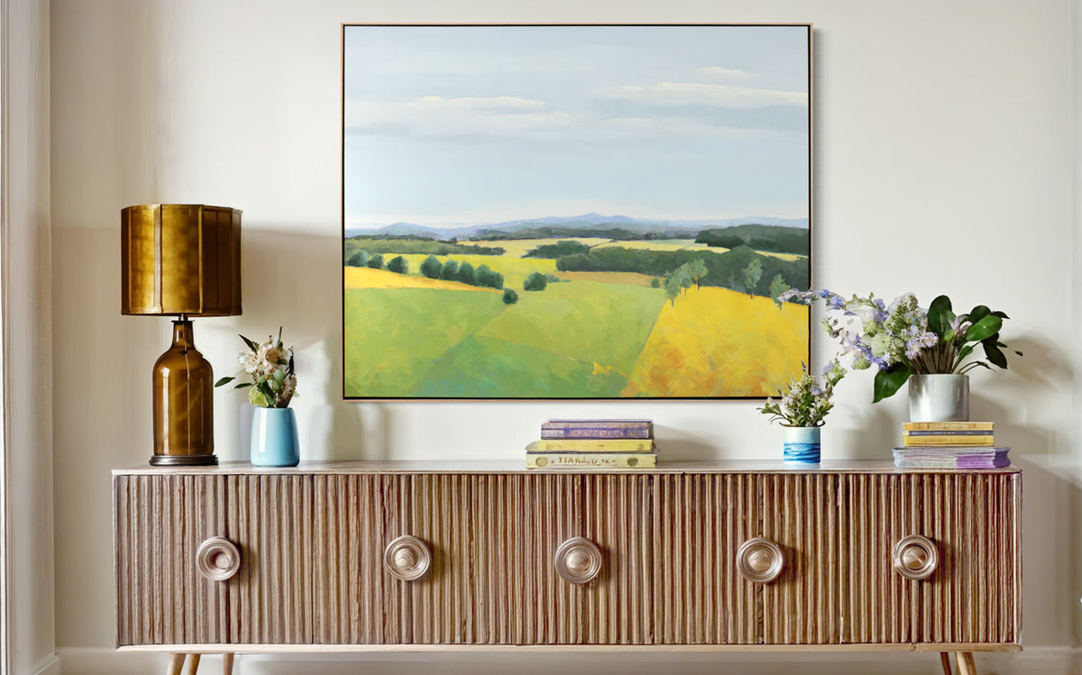 A blue and yellow landscape painting of a field hangs on a white wall above a wide console table with colorful table decor and a copper table lamp. 