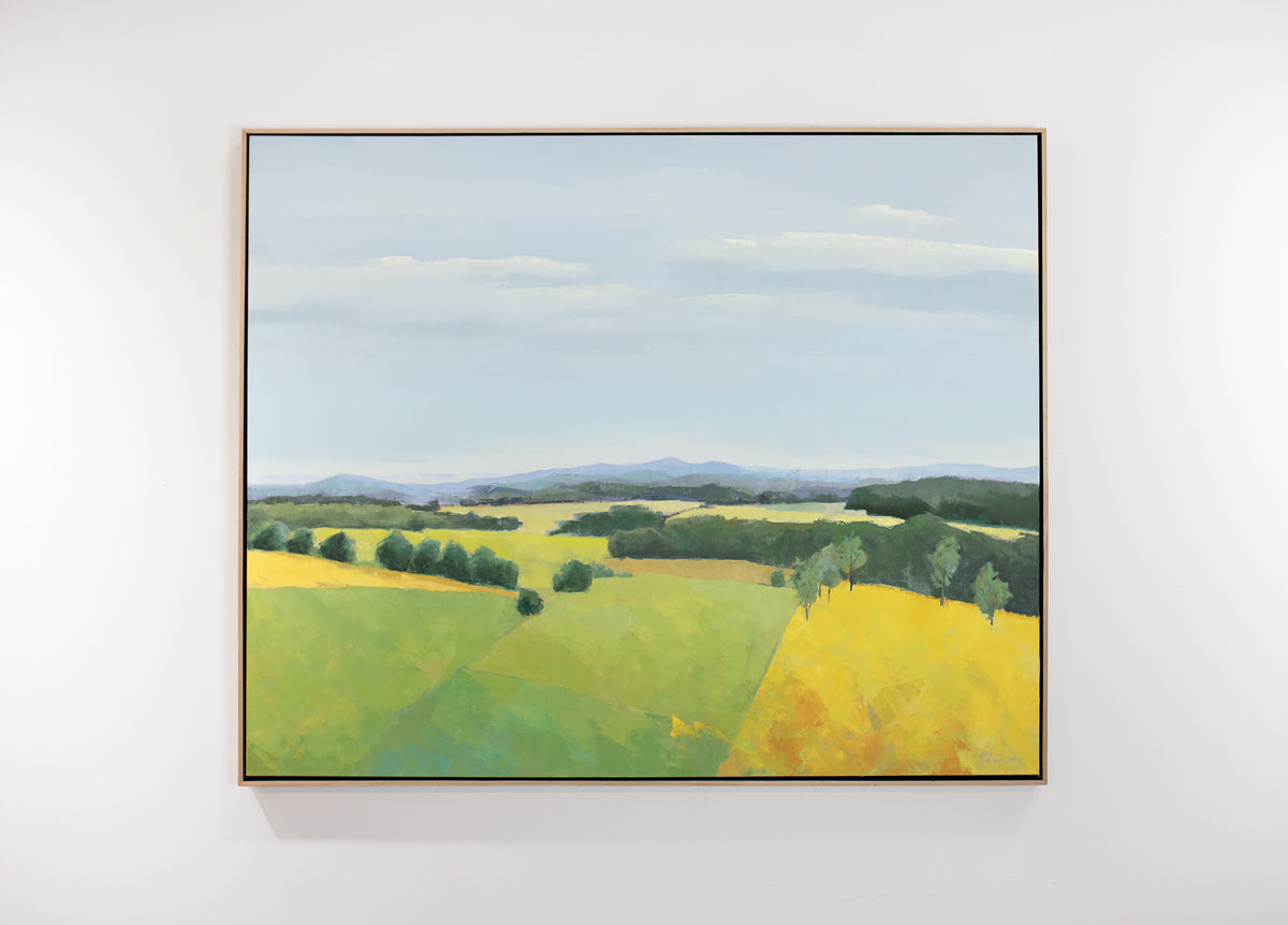 A framed blue and yellow landscape painting of a field hangs on a white wall.