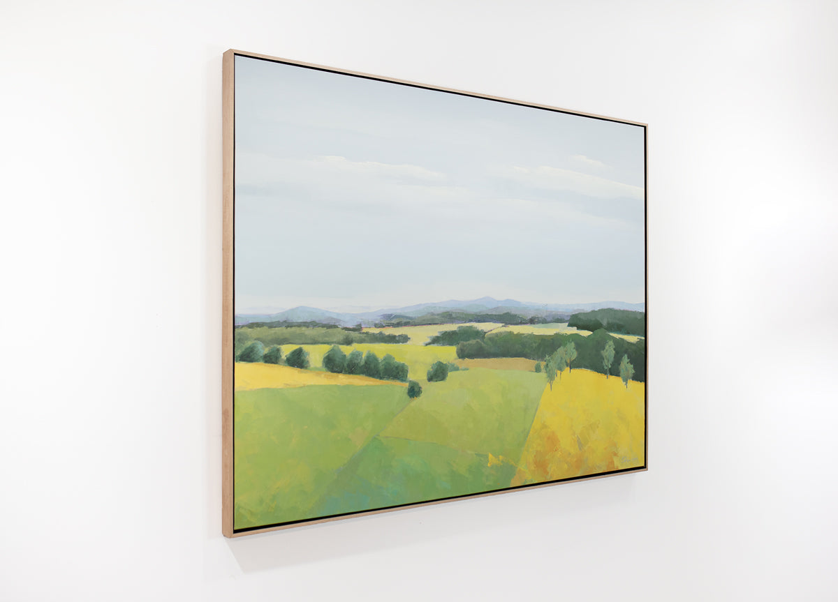 A framed blue and yellow landscape painting of a field hangs on a white wall.