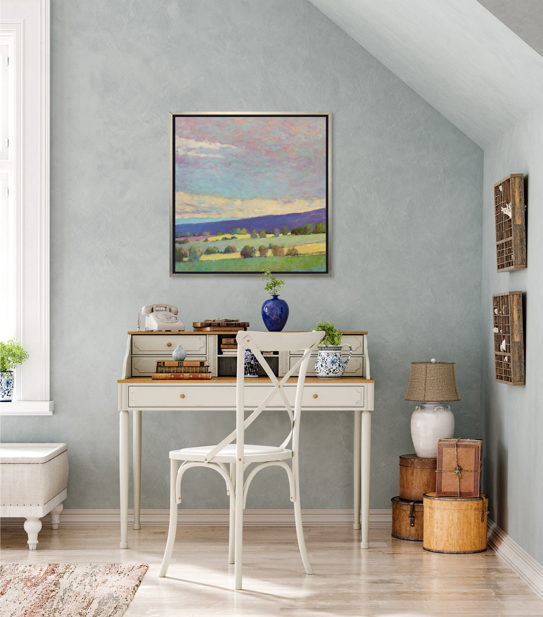 A multicolor abstract landscape print in a silver floater frame hangs on a light teal textured wall above a white two-tiered desk with accessories, a white chair and a stack of hat boxes in the corner.
