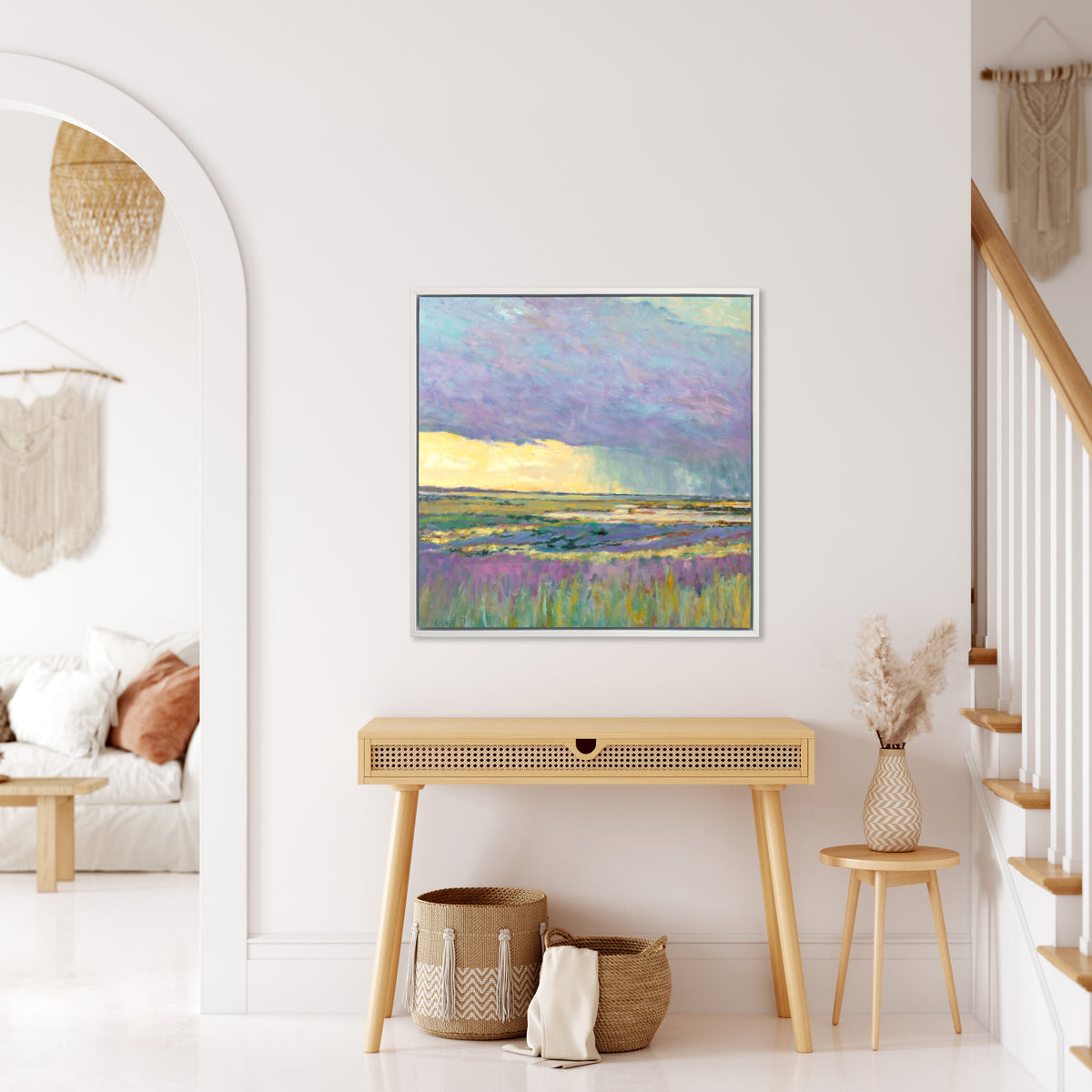 A multicolored abstract landscape print in a white floater frame hangs on a white wall above a wooden console table, a small round wooden side table with a vase and flowers and two woven baskets on the floor below.