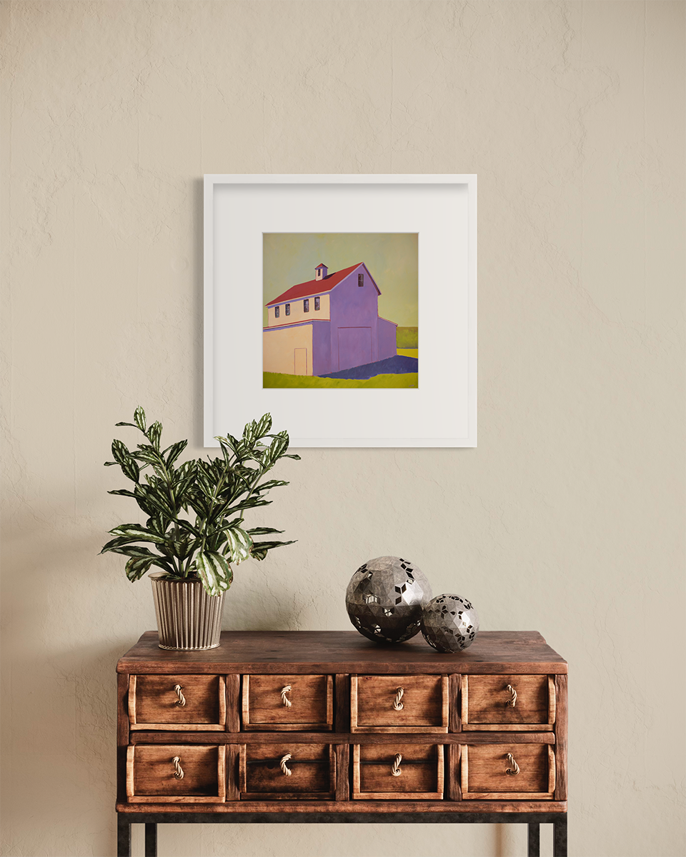 A contemporary barn print in a white frame with a mat hangs on a beige wall above a wood side table with a potted plant and two hammered steel spheres.