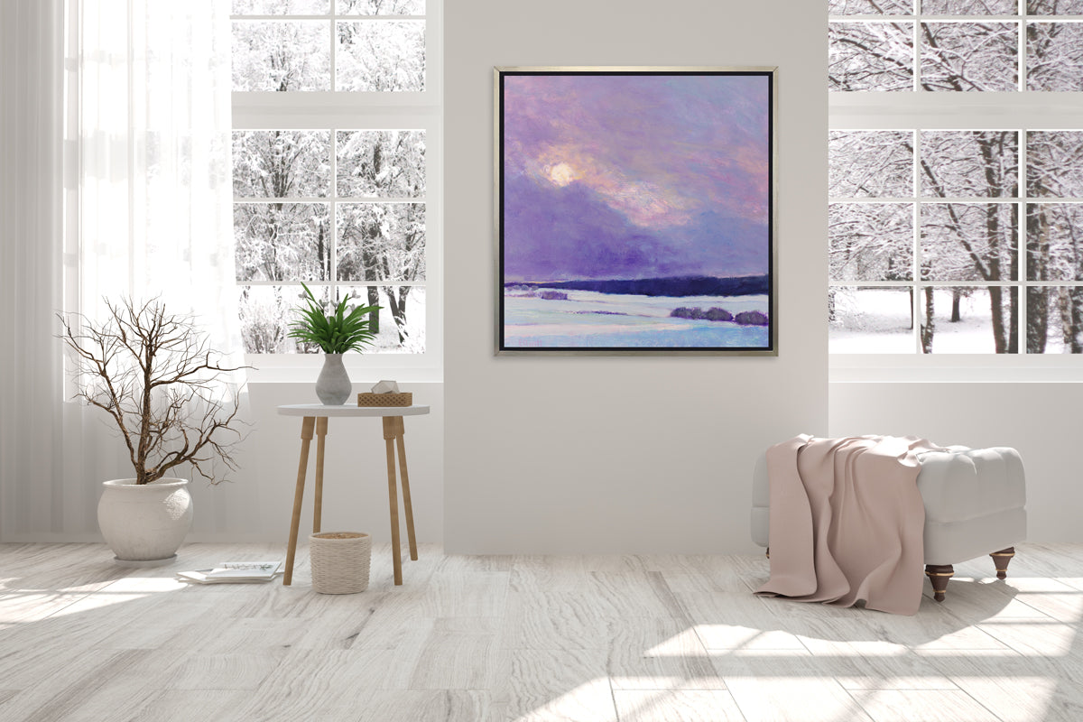 A purple and white snow landscape in a silver floater frame hangs on a white wall in between two large windows, a white tufted ottoman with a blush throw blanket and a side table with a vase with greenery.
